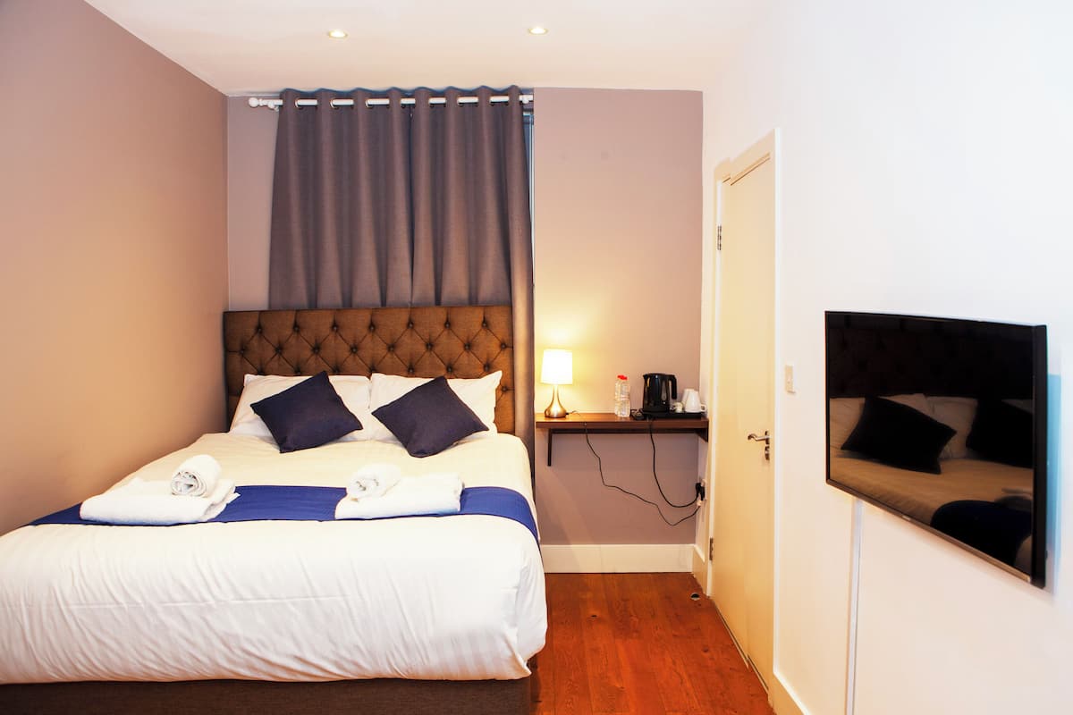 Una habitación de hotel con una cama grande, una mesita de noche con lámpara, un televisor de pantalla plana en la pared y una puerta.