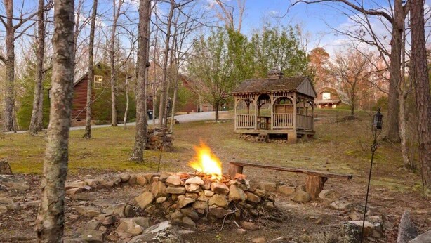 A rustic outdoor setting with a stone fire pit, a wooden gazebo, and a cabin in the background. - Scenic Solitude at Cabins in the Clouds! (Rising Fawn)