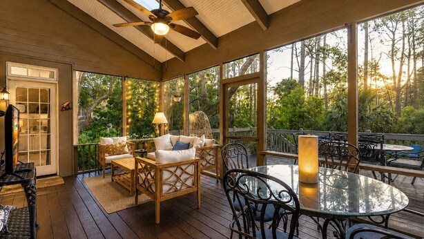 Eine überdachte Terrasse im Freien mit Holzboden, Deckenventilator und einem Glastisch, umgeben von Stühlen.