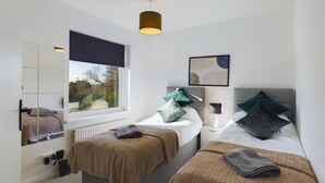 Ein modernes Schlafzimmer mit zwei Betten, einem großen Fenster, einer Deckenleuchte und einem gerahmten Bild an der Wand.