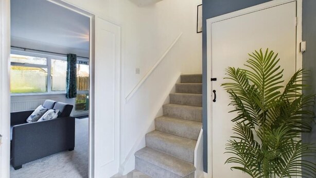 Eine Treppe führt zu einer Empfangshalle mit einer Tür, einem Blumentopf und Blick auf einen Raum mit Sofa und Fenster.