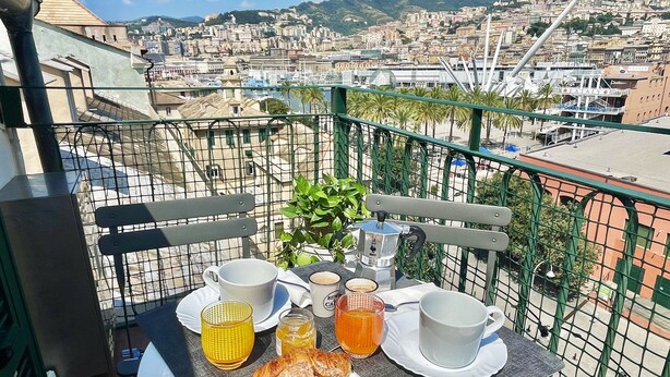 A balcony with a table set for breakfast, including croissants, coffee, and juice, overlooking a cityscape with buildings and a hill. - Happy Family Apartments Panorama (Genoa)