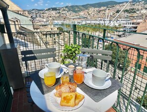 A balcony with a table set for breakfast, including croissants, coffee, and juice, overlooking a cityscape with buildings and a hill. - Happy Family Apartments Panorama (Genoa)