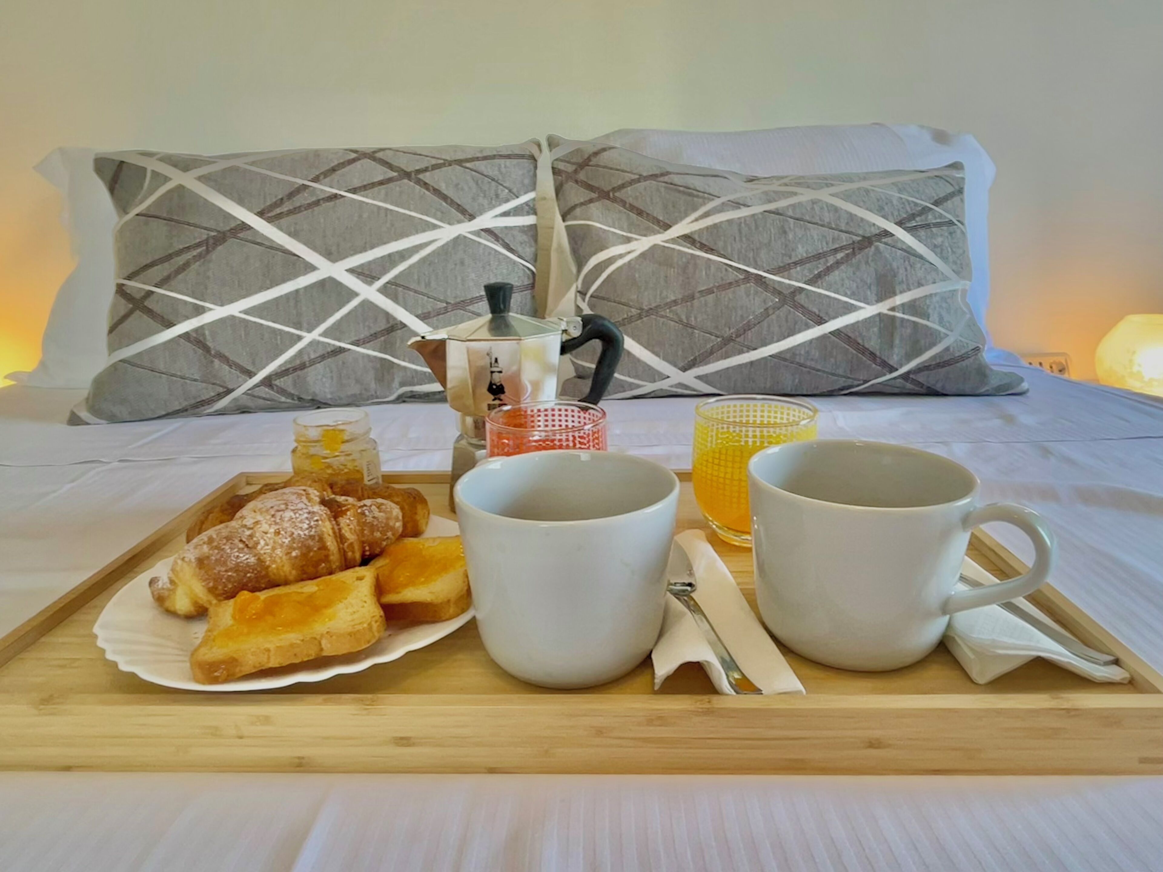 A breakfast tray with pastries, two mugs, and a coffee pot on a bed with pillows and a night lamp.