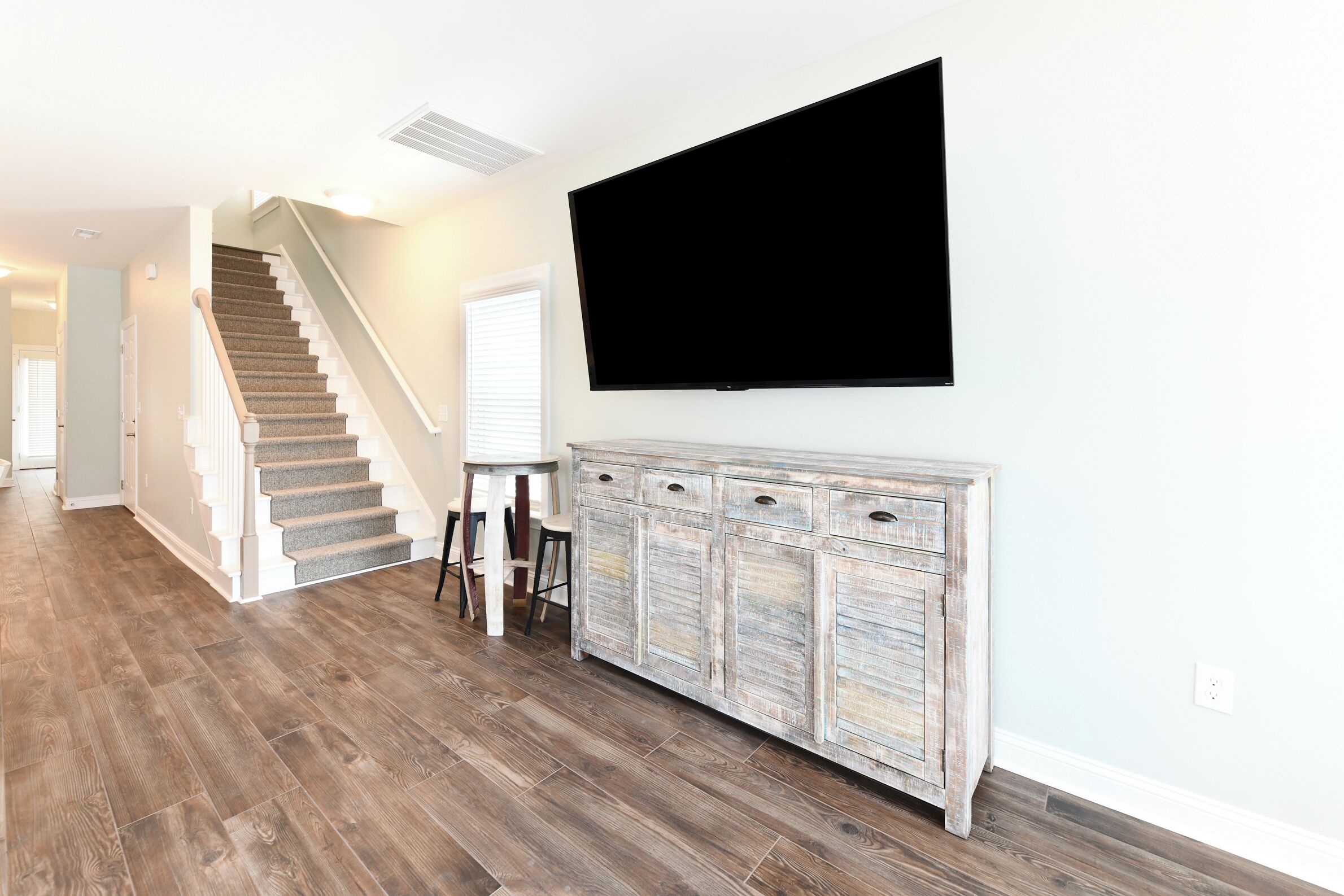 A spacious living area with a staircase, a large flat-screen TV mounted on the wall, and a rustic wooden cabinet.
