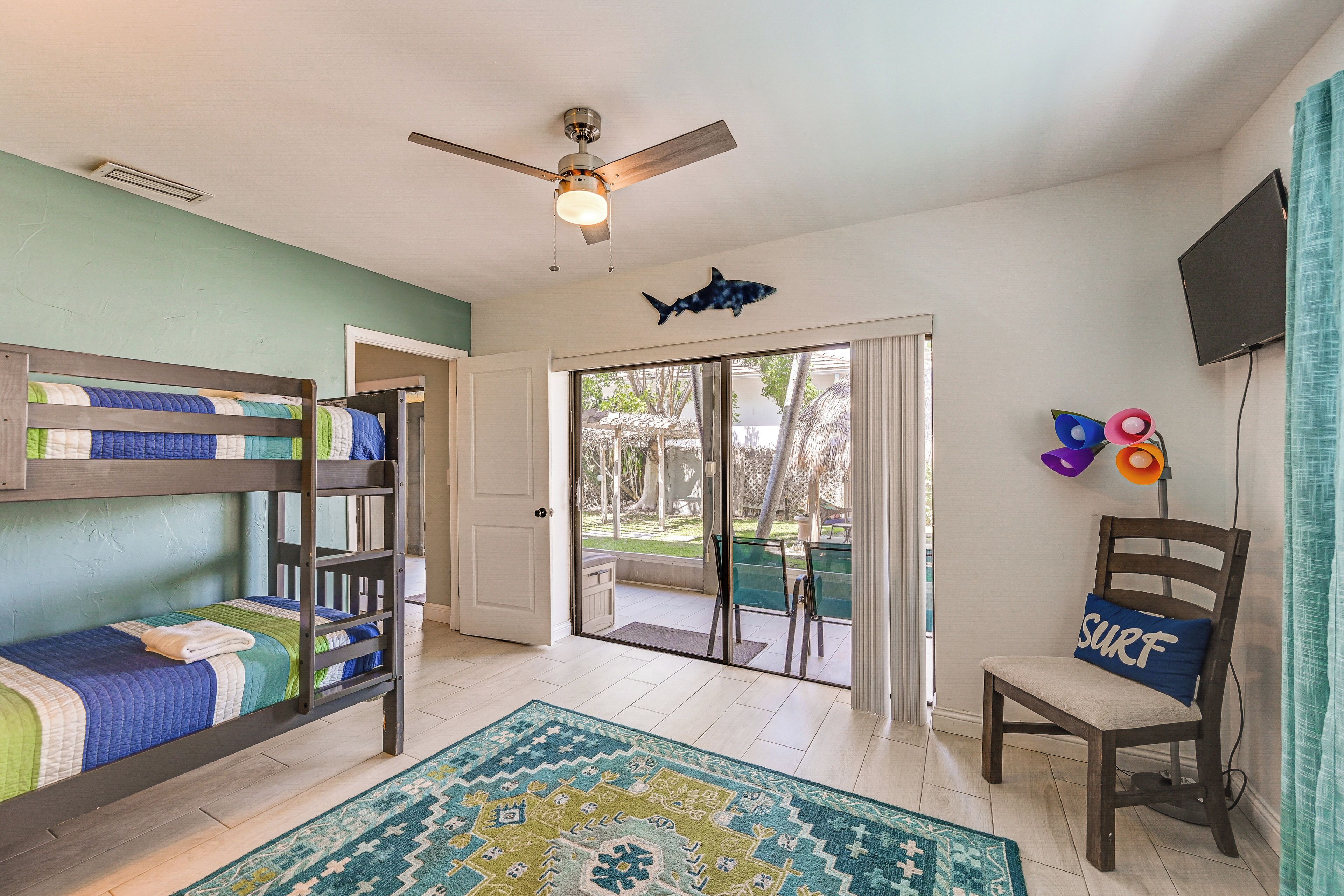 A room with bunk beds, a ceiling fan, a shark decoration, a surf-themed pillow, and a view to a patio.