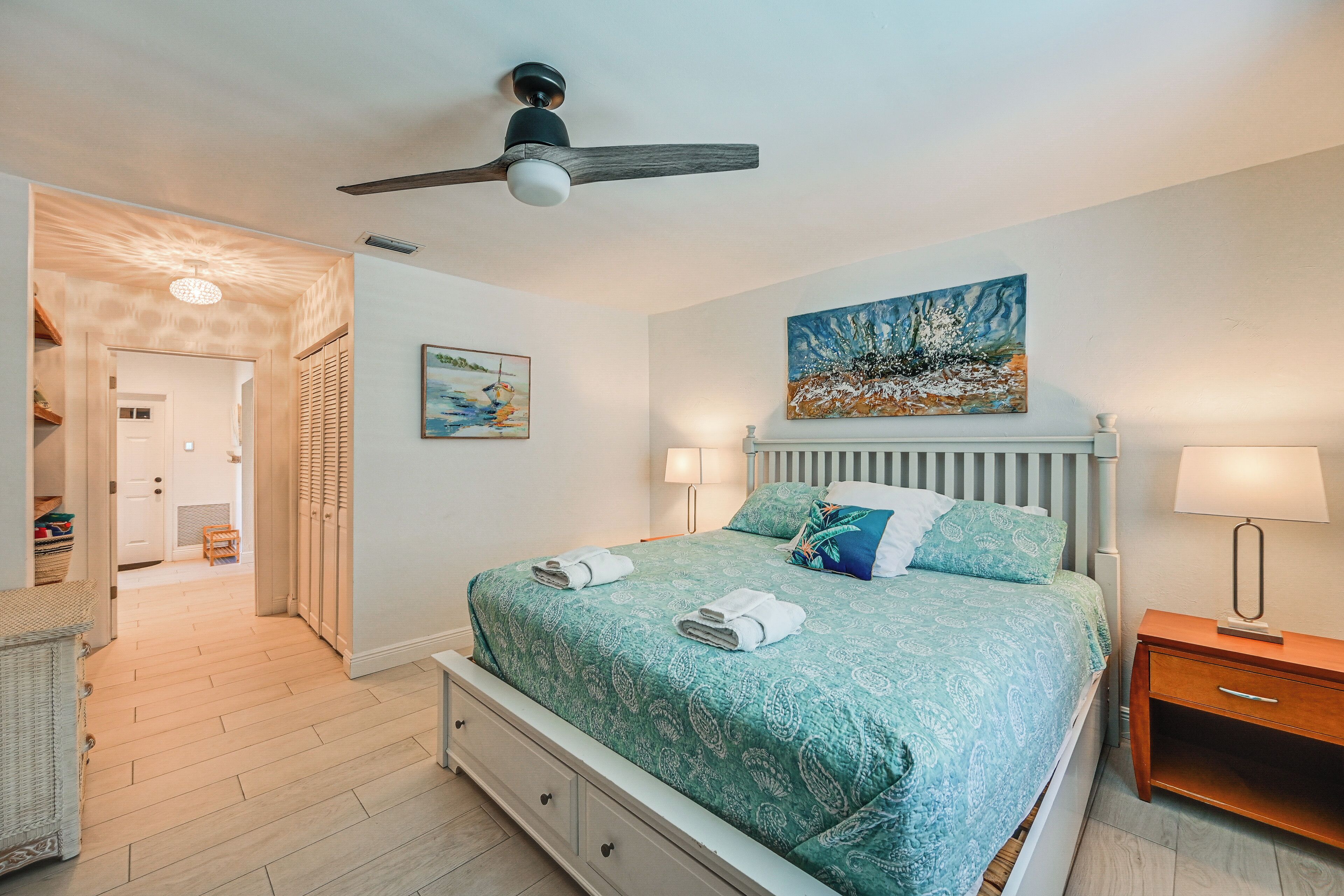 Un dormitorio con una cama, ventilador de techo, un cuadro, mesita de noche y lámpara.