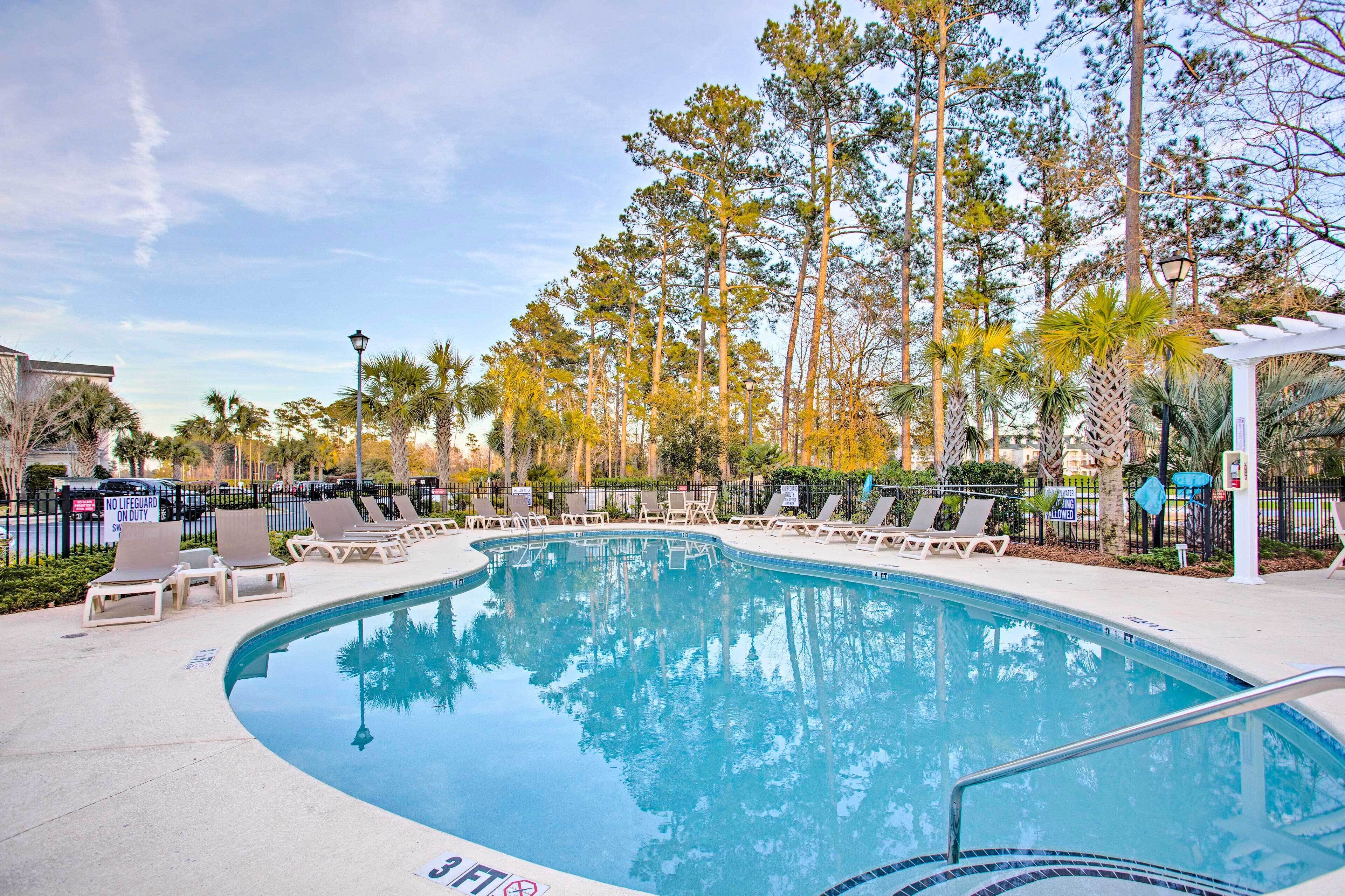 A swimming pool with lounge chairs and a pergola, surrounded by tall trees and a clear sky.