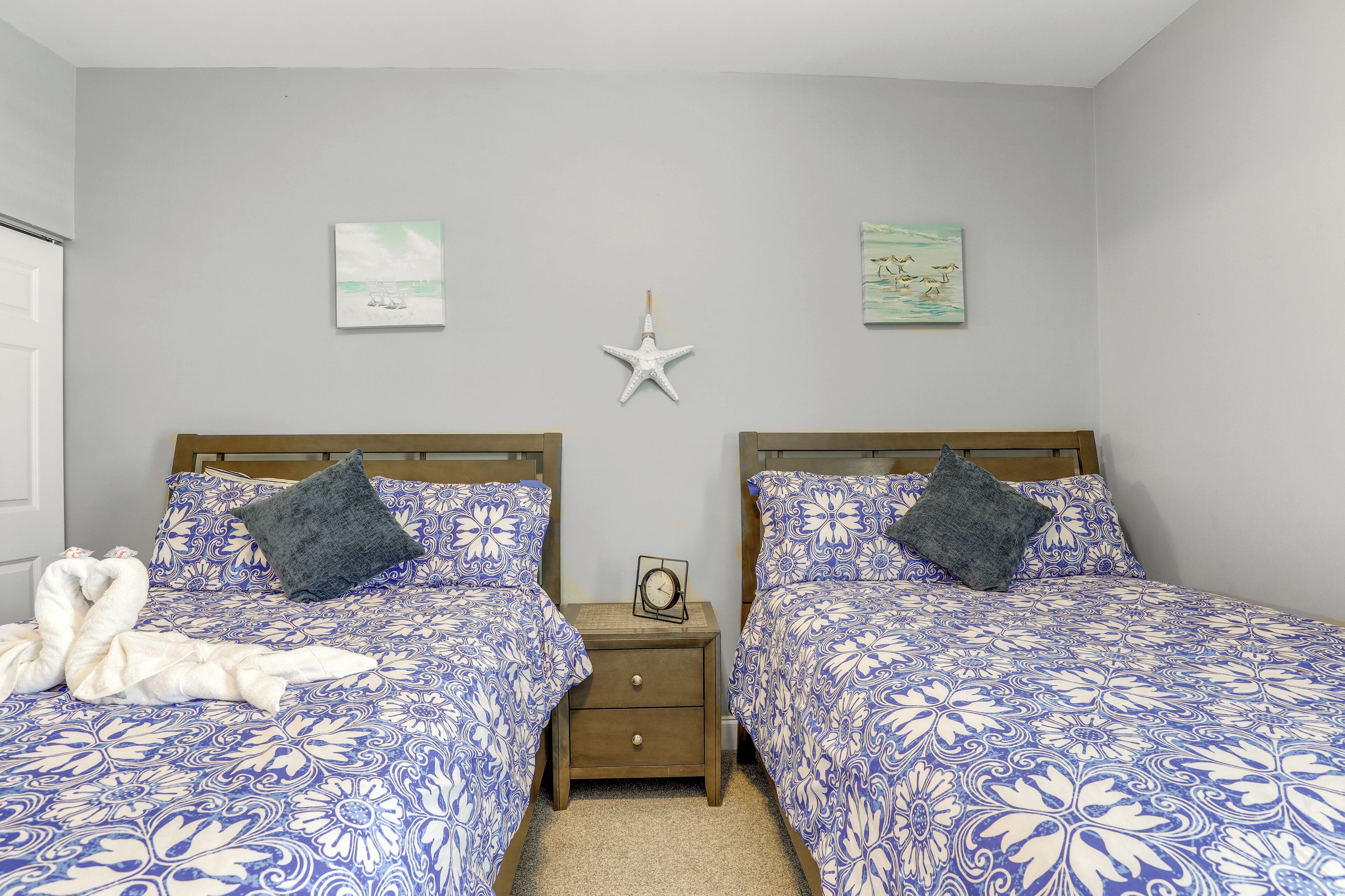 Ein Doppelzimmer mit zwei Betten, jeweils mit einer blauen, floralen Tagesdecke, einem Holz-Kopfstück und einem dunkelgrauen Kopfkissen. Zwischen den Betten steht ein Nachttisch mit einer Uhr, und an der Wand ist eine Stern-Dekoration angebracht.