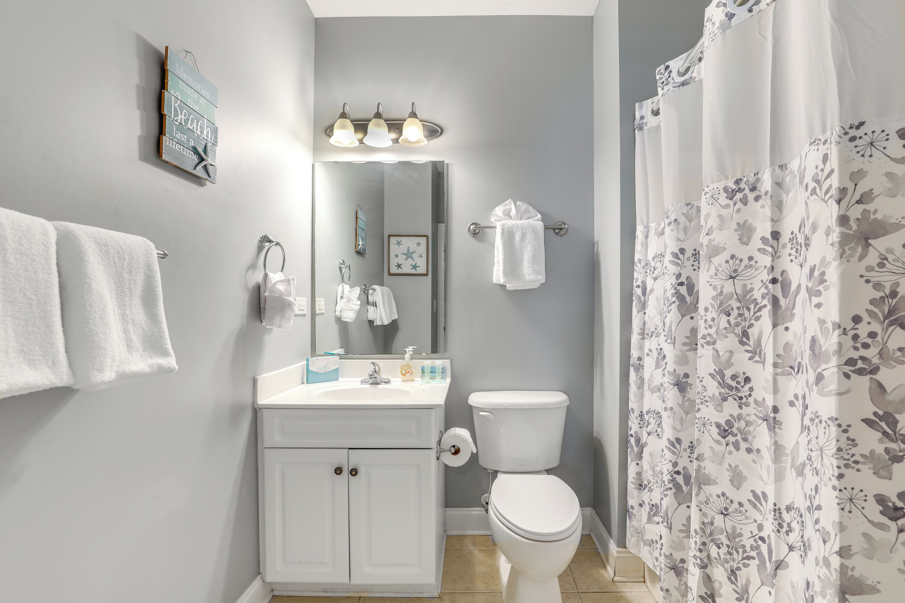 Un baño con tocador blanco, inodoro, lavamanos con espejo y cortina de ducha estampada con motivos florales.