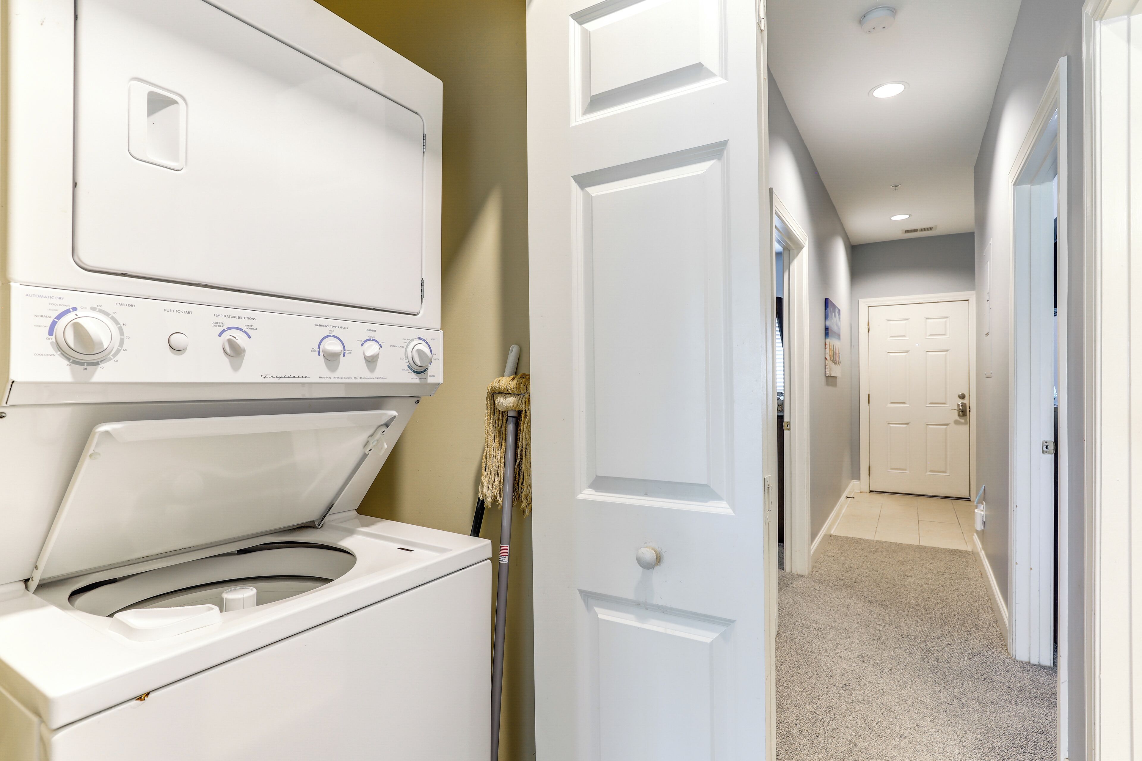 A laundry room with a front-loading washer and dryer, a mop, and a hallway leading to a door.
