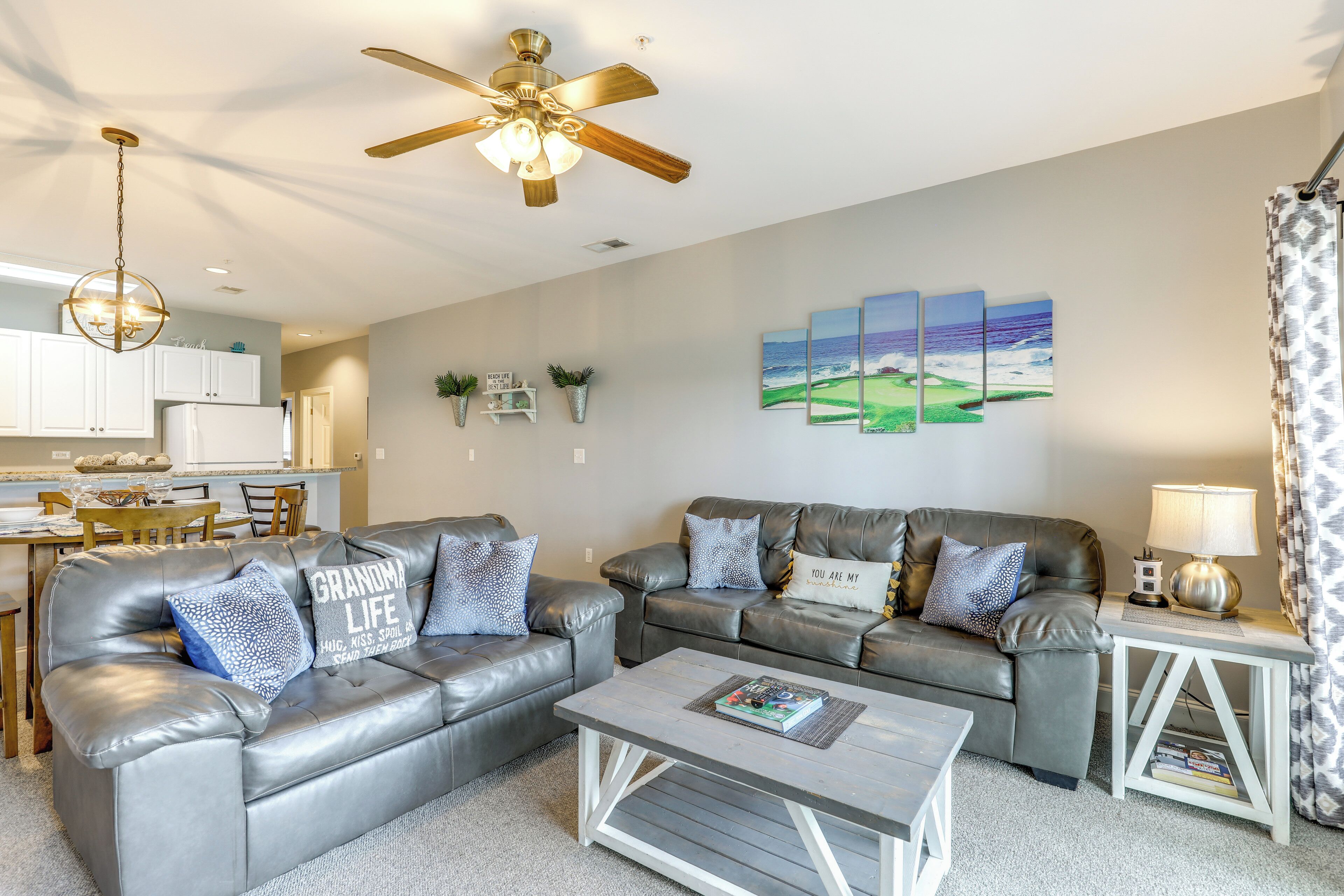 A modern living room with two grey leather sofas, a coffee table, a ceiling fan, and a kitchen in the background.