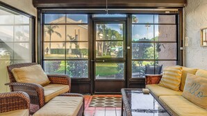 A room with wicker furniture, a glass table, and a view of a garden through large windows. - Pool, Golf & Beach Access: Fort Pierce Condo! (Fort Pierce)
