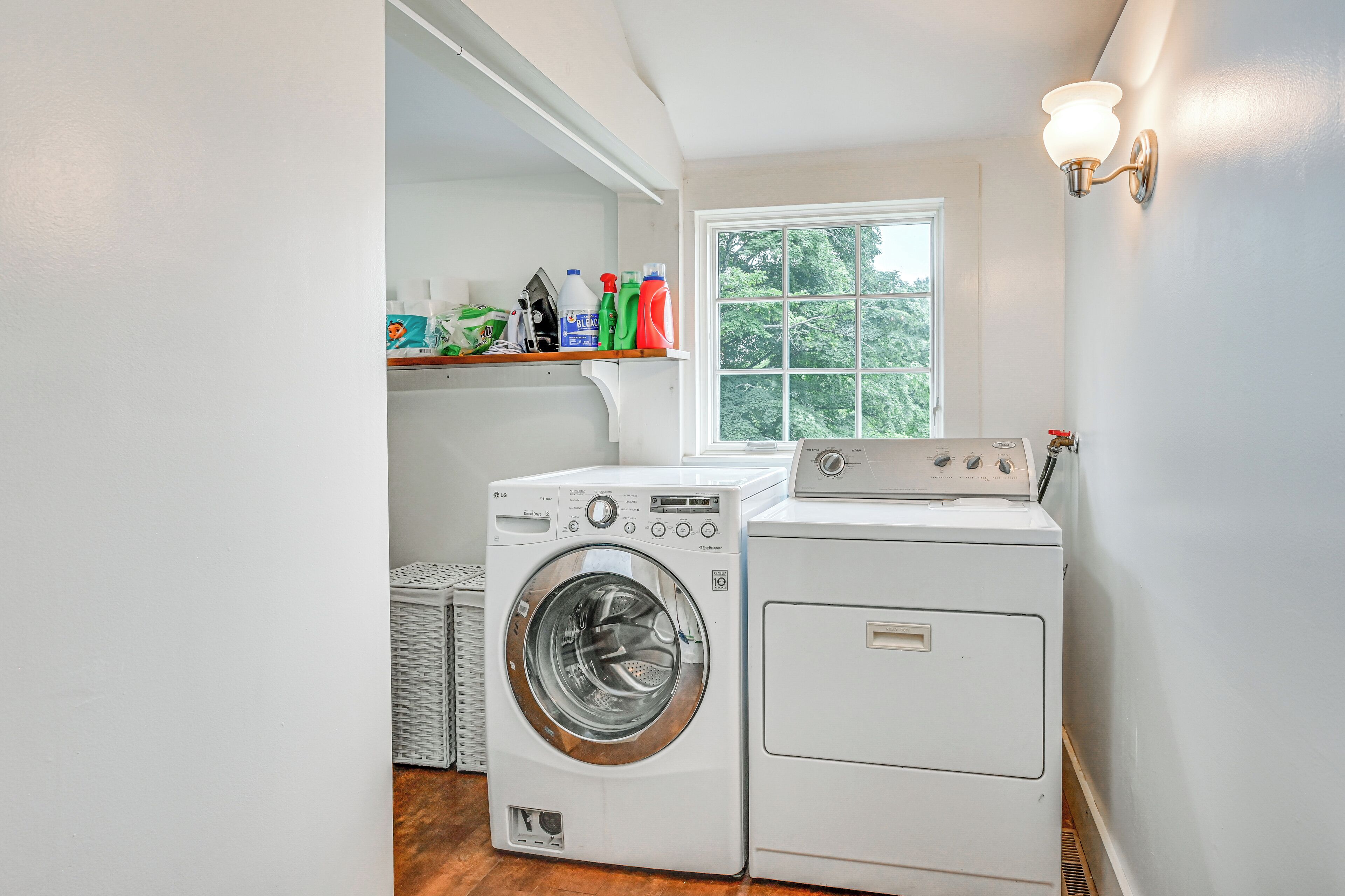 A laundry room with two washing machines, a shelf with cleaning supplies, and a window with a view of trees.
