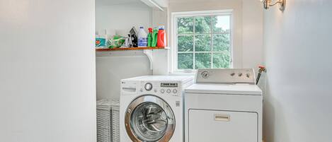 A laundry room with two washing machines, a shelf with cleaning supplies, and a window with a view of trees.