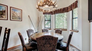 Une salle à manger avec une table ronde, six chaises à motifs, un lustre orné de bois de cerf, deux tableaux encadrés et de grandes fenêtres donnant sur la forêt.