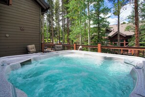 Un bain à remous entouré d’une terrasse en bois, avec vue sur une zone boisée et un bâtiment en arrière-plan.