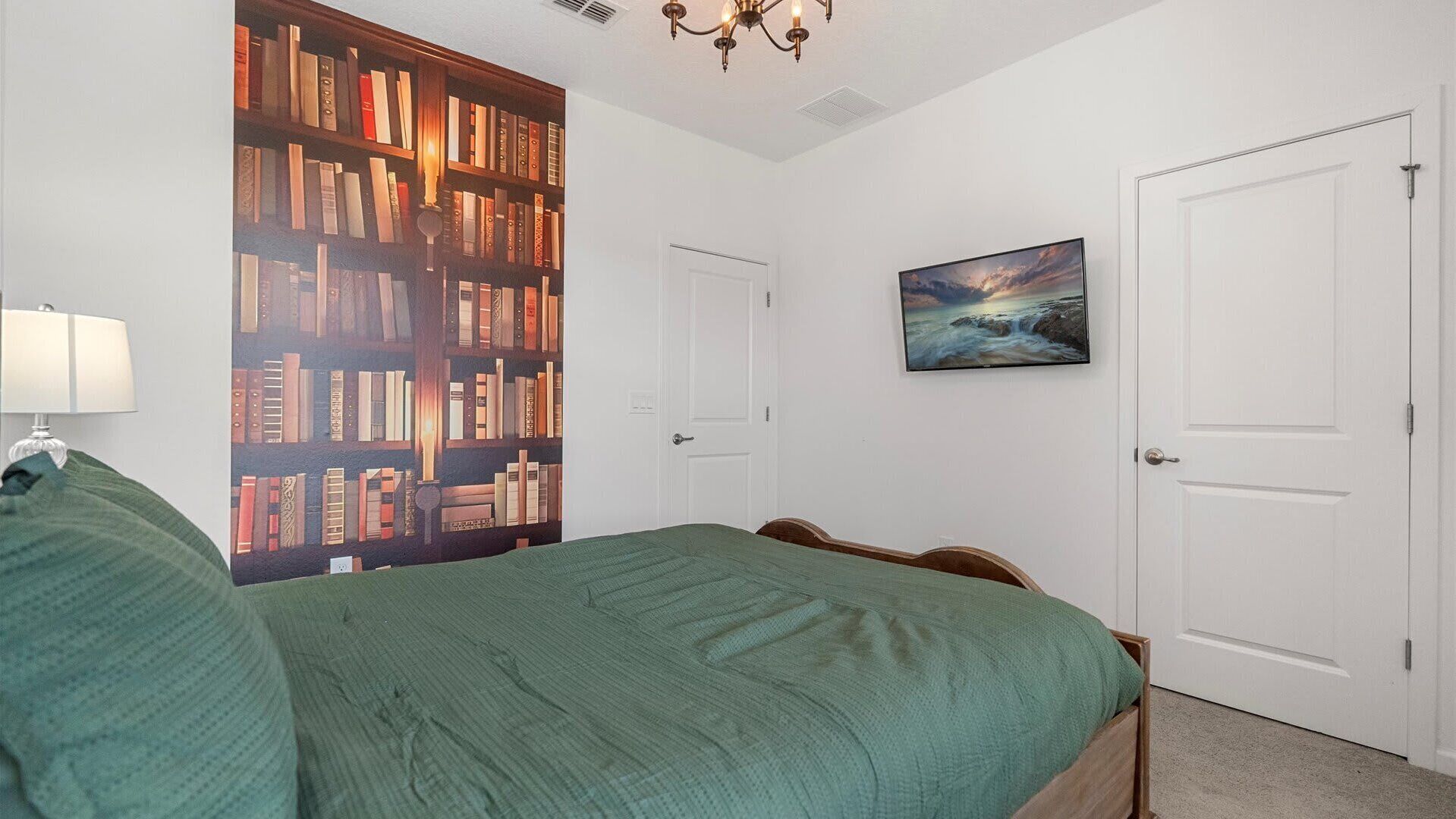 A bedroom with a large bookshelf, a bed with a green blanket, a painting of a seascape, and white doors.