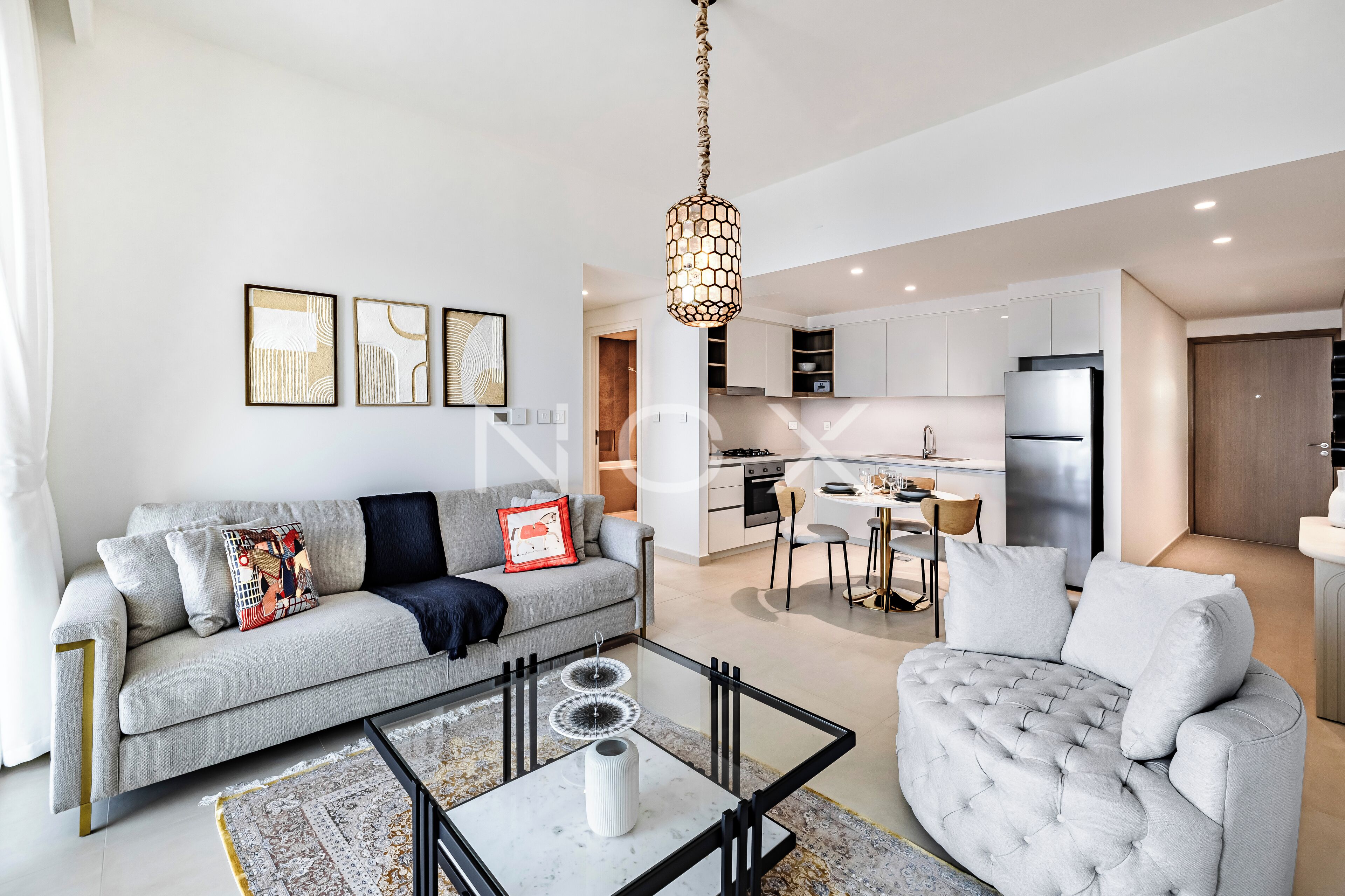 A modern living room with a grey sofa, a glass coffee table, and a dining area with a kitchen in the background.