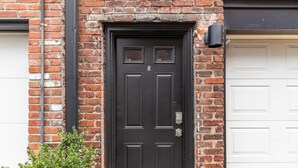 Een zwarte deur met het cijfer '5', geplaatst in een bakstenen muur, met een witte garagepoort ernaast.