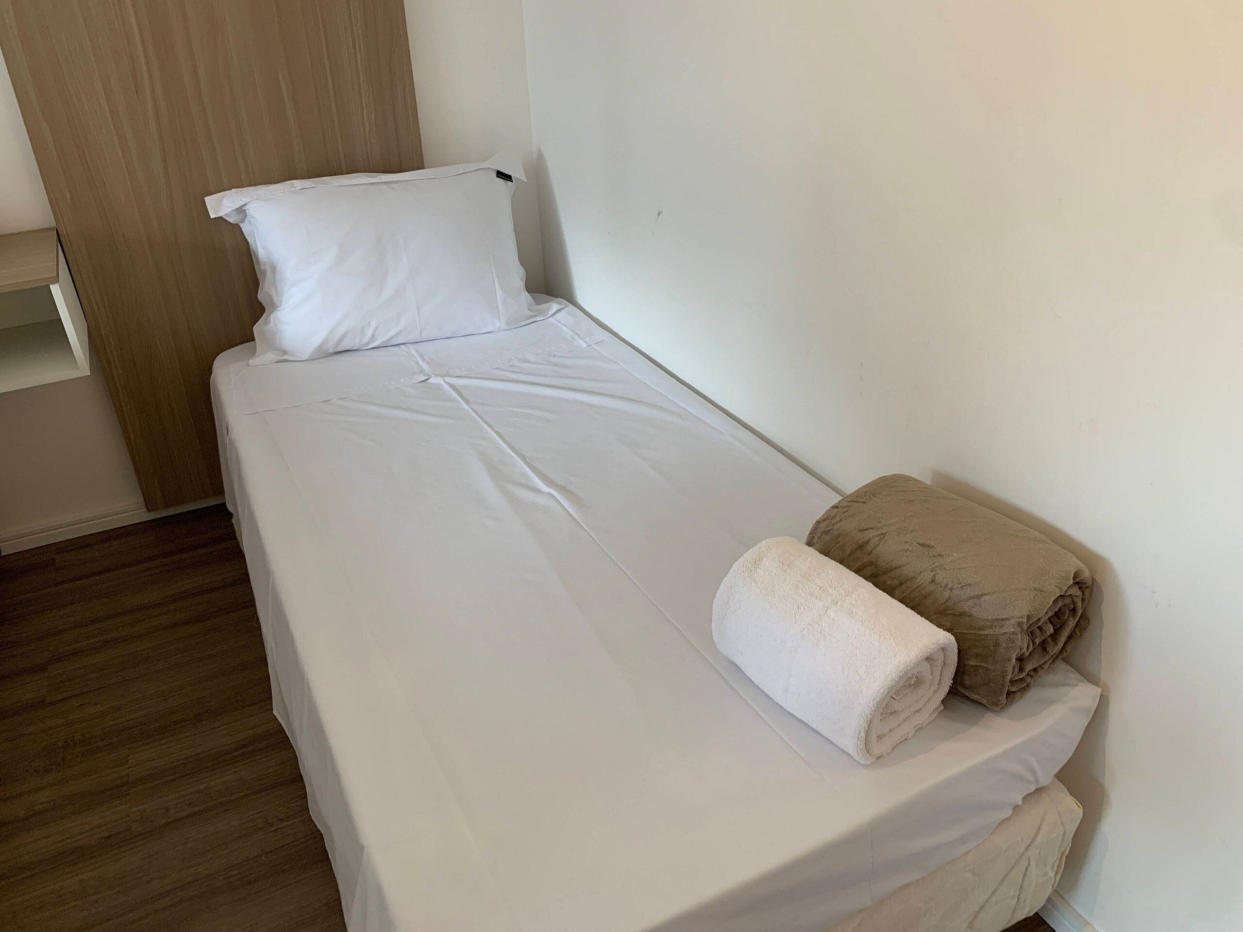 A single bed with white bedding and a rolled towel, a wooden panel wall, and a wooden floor.