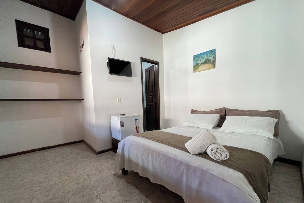 A bedroom with a bed, a small white refrigerator, a wall-mounted TV, and a door.