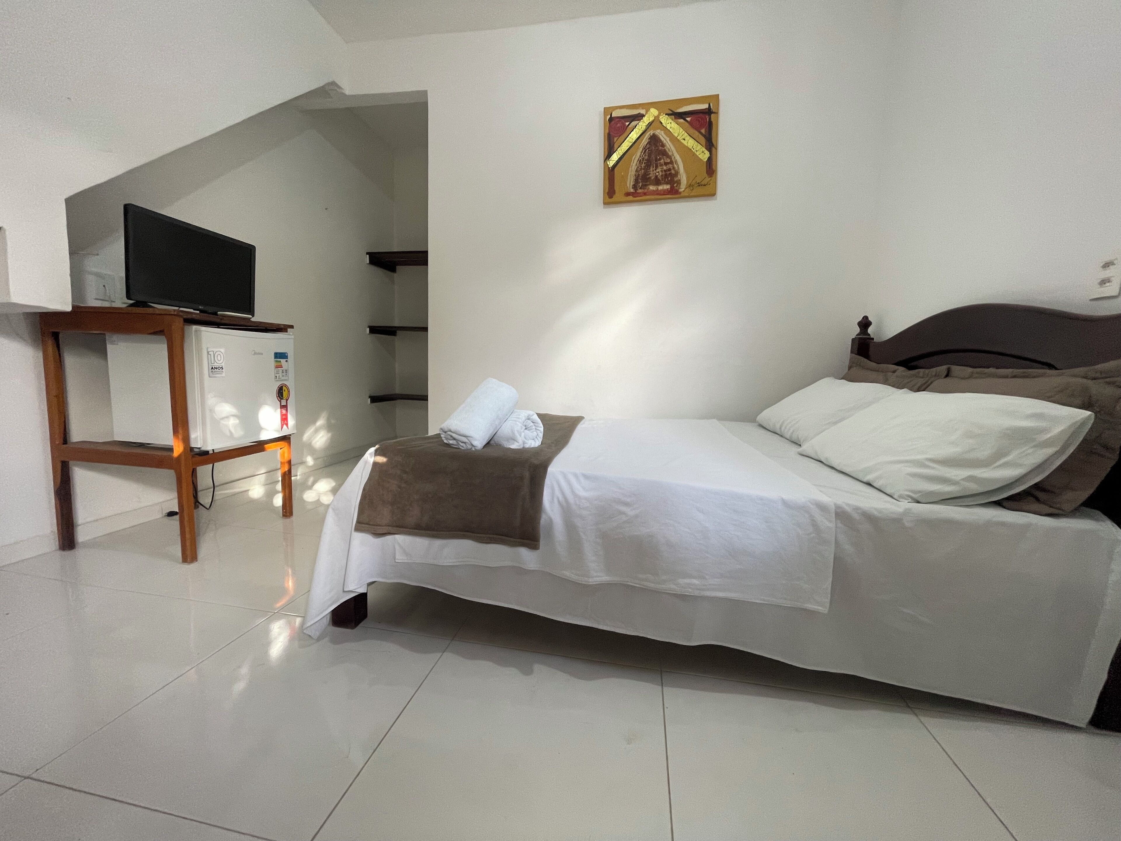 A bedroom with a bed, a TV on a stand, a small table, and a painting on the wall.