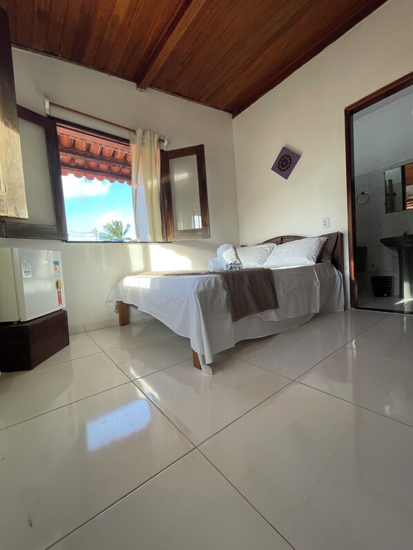 A bedroom with a bed, a window with curtains, a mirror, and a small kitchenette area.