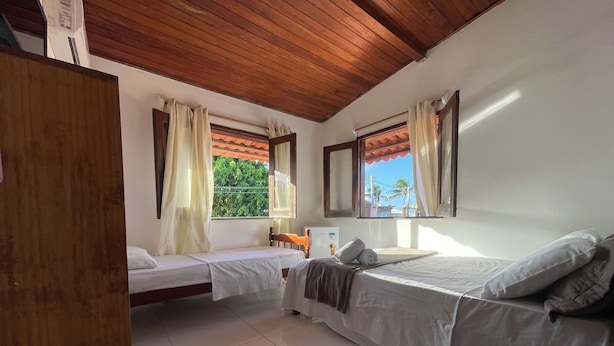 A room with two beds, a wooden ceiling, a window with a view of greenery, and a tiled floor. - Pousada Vila Nova (Cairu)