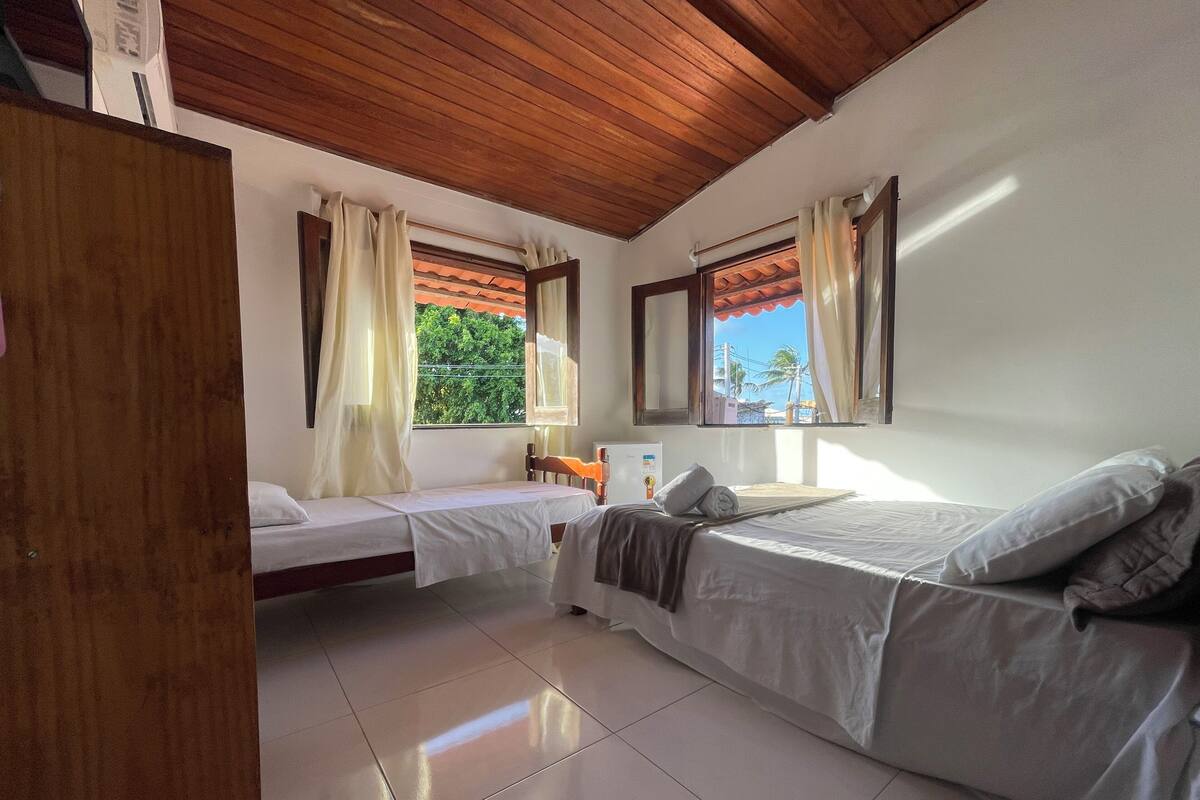 A room with two beds, a wooden ceiling, a window with a view of greenery, and a tiled floor.