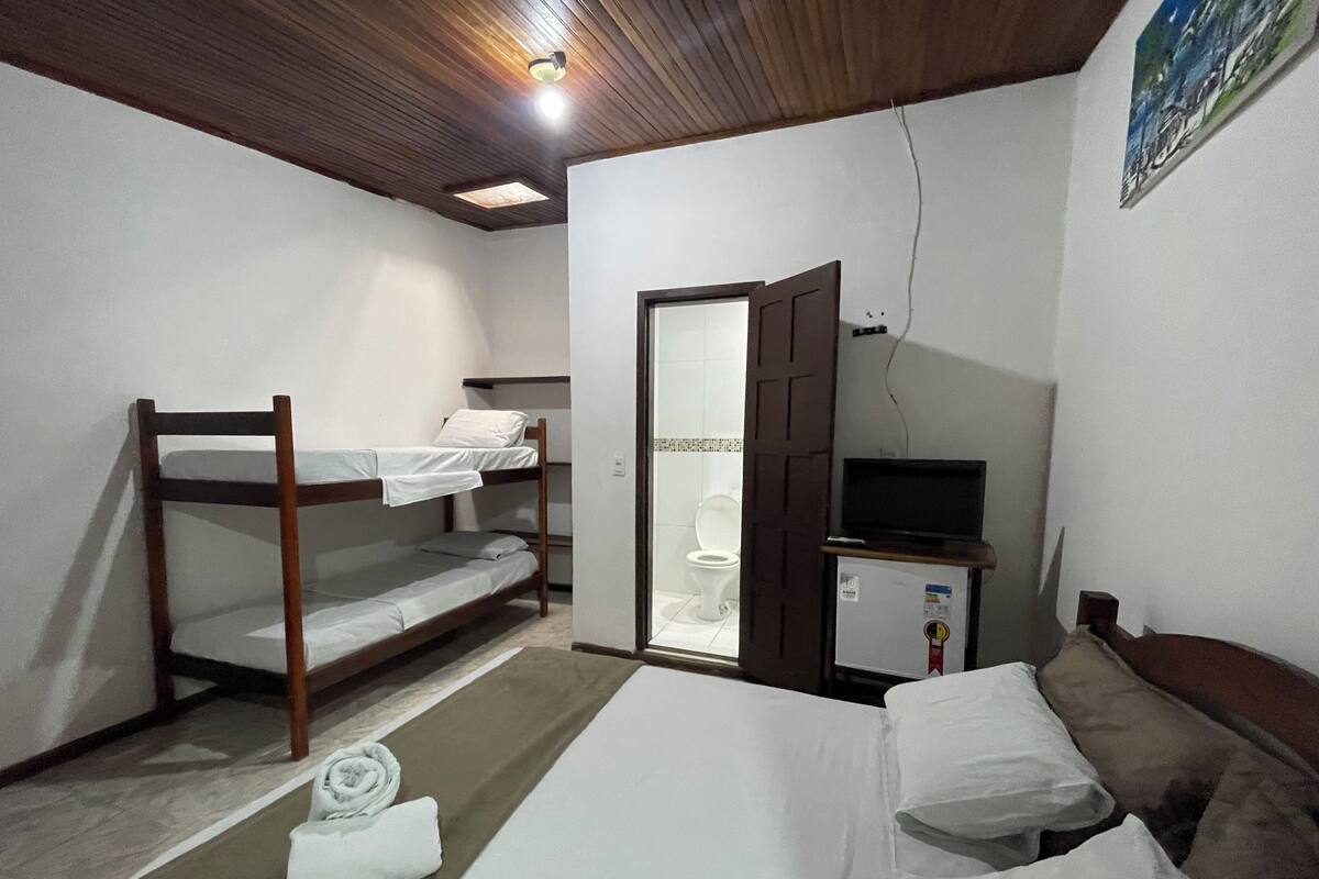 A small, compact room with bunk beds, a single bed, a toilet, and a small cabinet with a television.
