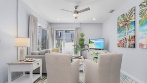 A modern living room with a sofa, armchairs, a TV, a ceiling fan, and a large window with curtains. - Dune Our Thing - Building A - 171 Washington 3 Bedroom Condo by RedAwning (Fort Myers Beach)