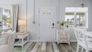 A room with a white door, a small table with a lamp, a white chair, and a window with a view of the outside. - Dune Our Thing - Building A - 171 Washington 3 Bedroom Condo by RedAwning (Fort Myers Beach)