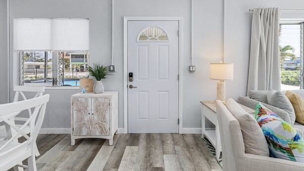 A modern living room with a white door, a window with blinds, a sofa with colorful pillows, and a small side table with a lamp. - Dune Our Thing - Building A - 171 Washington 3 Bedroom Condo by RedAwning (Fort Myers Beach)