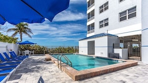A swimming pool with lounge chairs and umbrellas, a building with balconies, and a clear sky. - Beach Villas # 306 by RedAwning - 15% off through September 2024 (Fort Myers Beach)