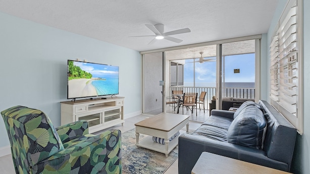 A living room with a television, sofa, coffee table, and patterned armchair. - Beach Villas # 306 by RedAwning - 15% off through September 2024 (Fort Myers Beach)