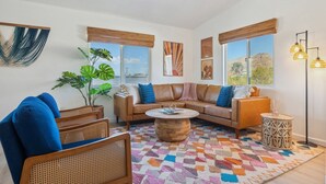 Un salon avec un canapé en cuir marron, des coussins bleus, une table basse en bois, des fauteuils en osier et un tapis coloré.