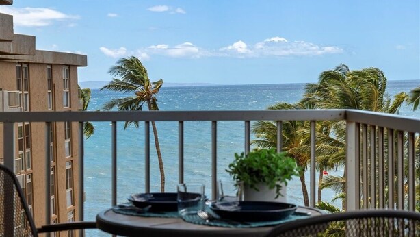 A balcony with a table set for dining, overlooking a beach with palm trees and the ocean. - Nani Kai Hale 604 1 Bedroom Condo (Kihei)