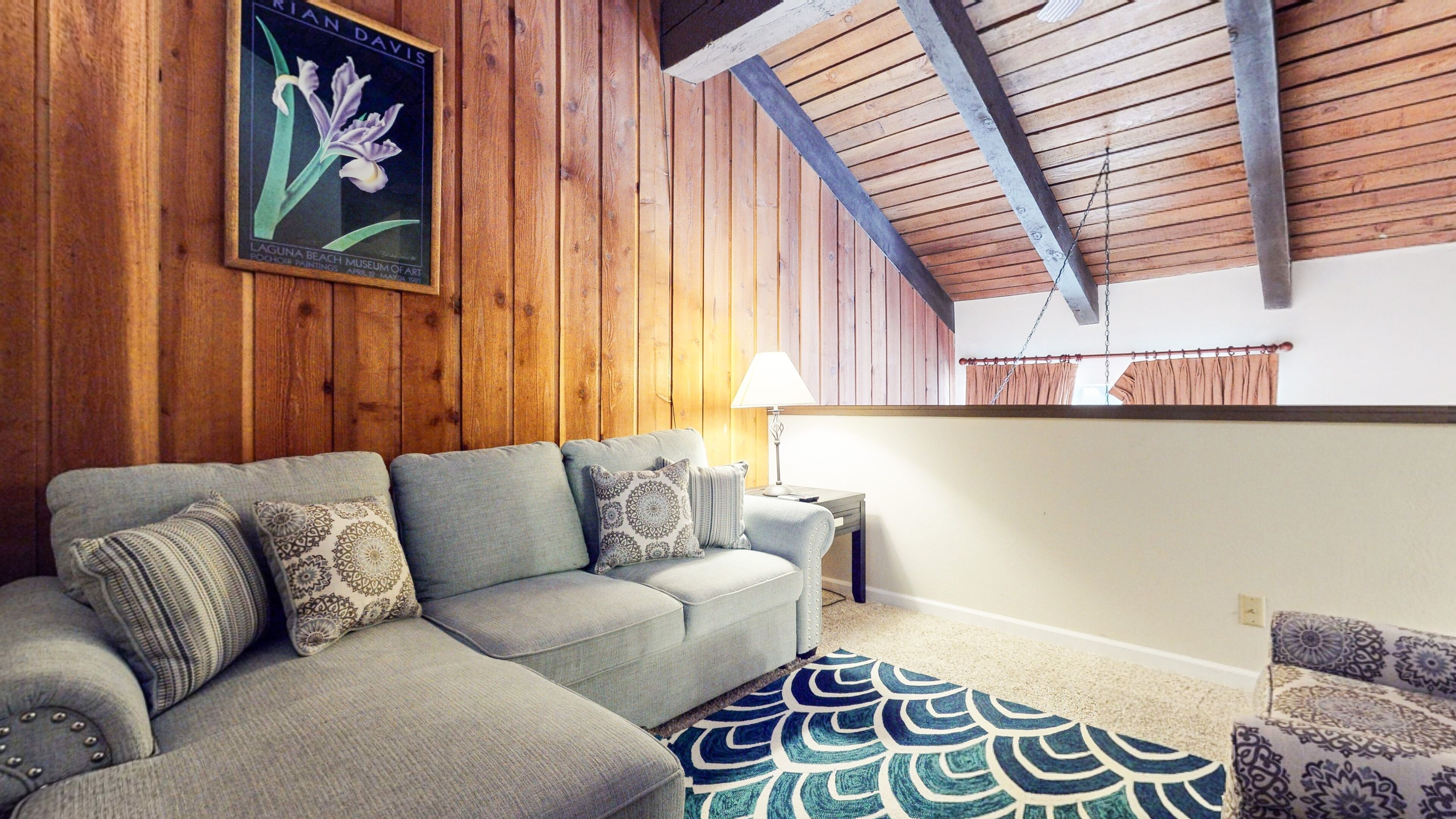 A living room with a grey sofa, patterned cushions, a blue and white rug, wooden walls, and a ceiling with exposed beams.
