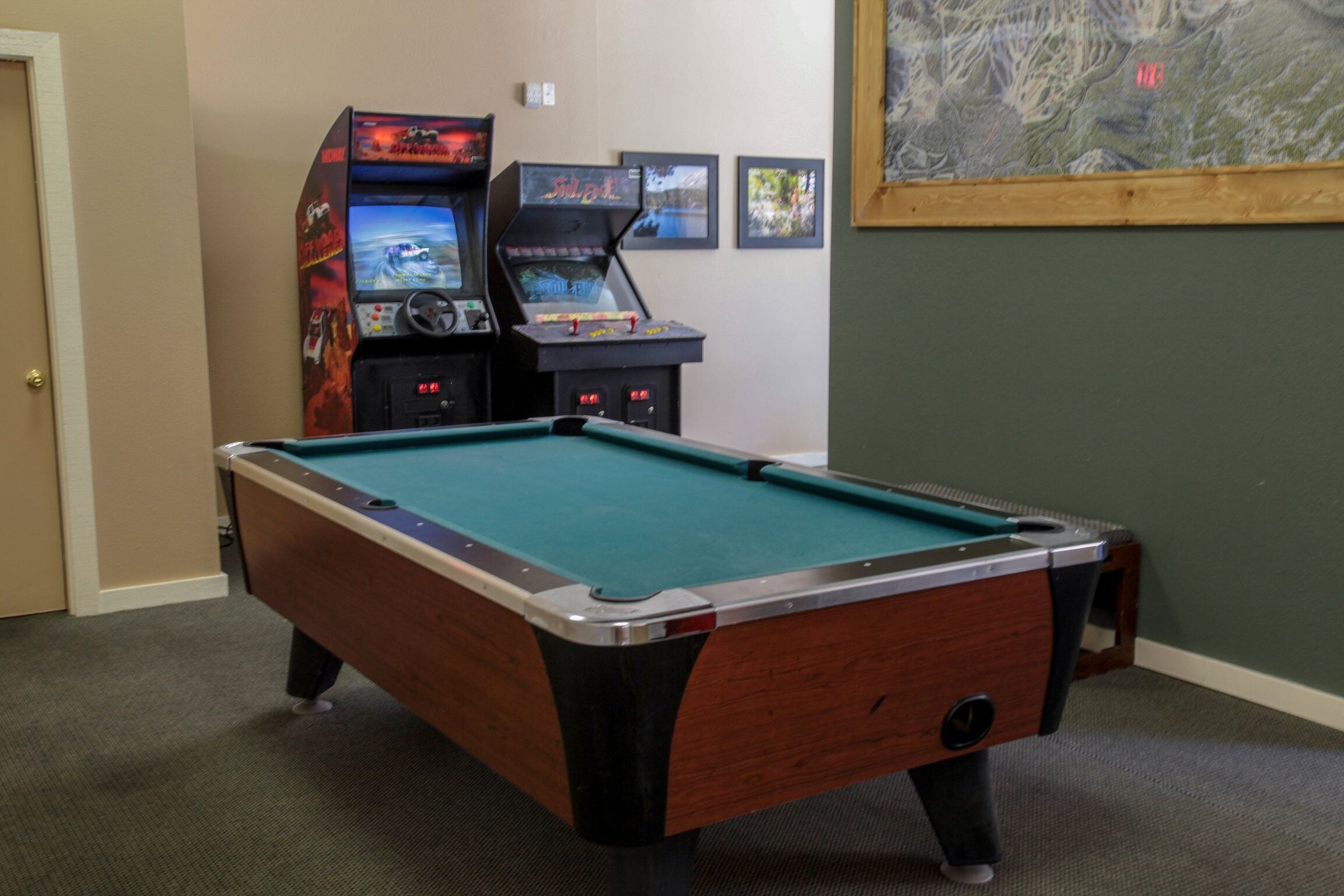 A pool table in a room with two arcade machines and a framed picture on the wall.
