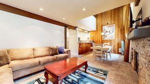 A living room with a sofa, coffee table, and patterned rug. There is a kitchen area with wooden cabinets and a dining table with chairs. A poster is visible on the wall.