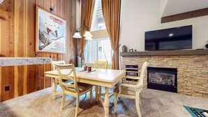 A cozy living room with a stone fireplace, wooden walls, a dining table with chairs, and a mounted TV.