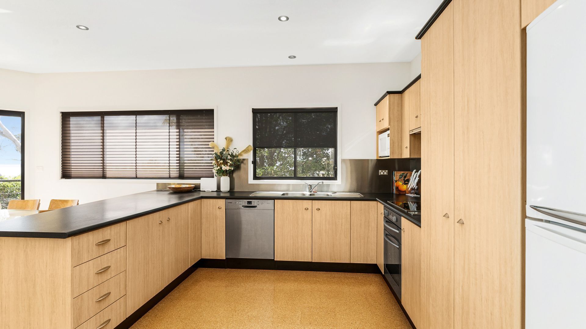A modern kitchen with wooden cabinets, a black countertop, and a dishwasher. There are two windows with blinds, and a refrigerator is visible on the right.