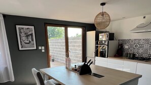 A modern kitchen with a central island, two bar stools, a hanging light fixture, and a view through the window. - Maison de Ville (Cambrai)