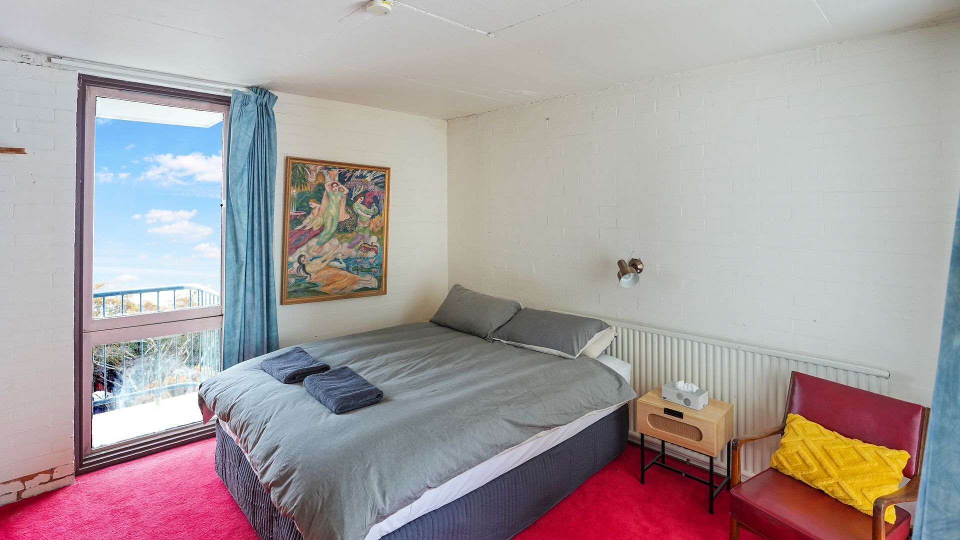 A bedroom with a large bed, a window with curtains, a painting on the wall, a small bedside table with a lamp, and a red chair.