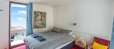 A bedroom with a large bed, a window with curtains, a painting on the wall, a small bedside table with a lamp, and a red chair.