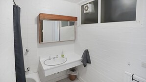 Un baño con un lavamanos blanco, un espejo con marco de madera, una toalla colgada en la pared y una ventana con rejilla.