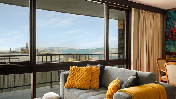 Una habitación con sofá gris, almohadones amarillos, vista a las montañas y un cuadro en la pared.