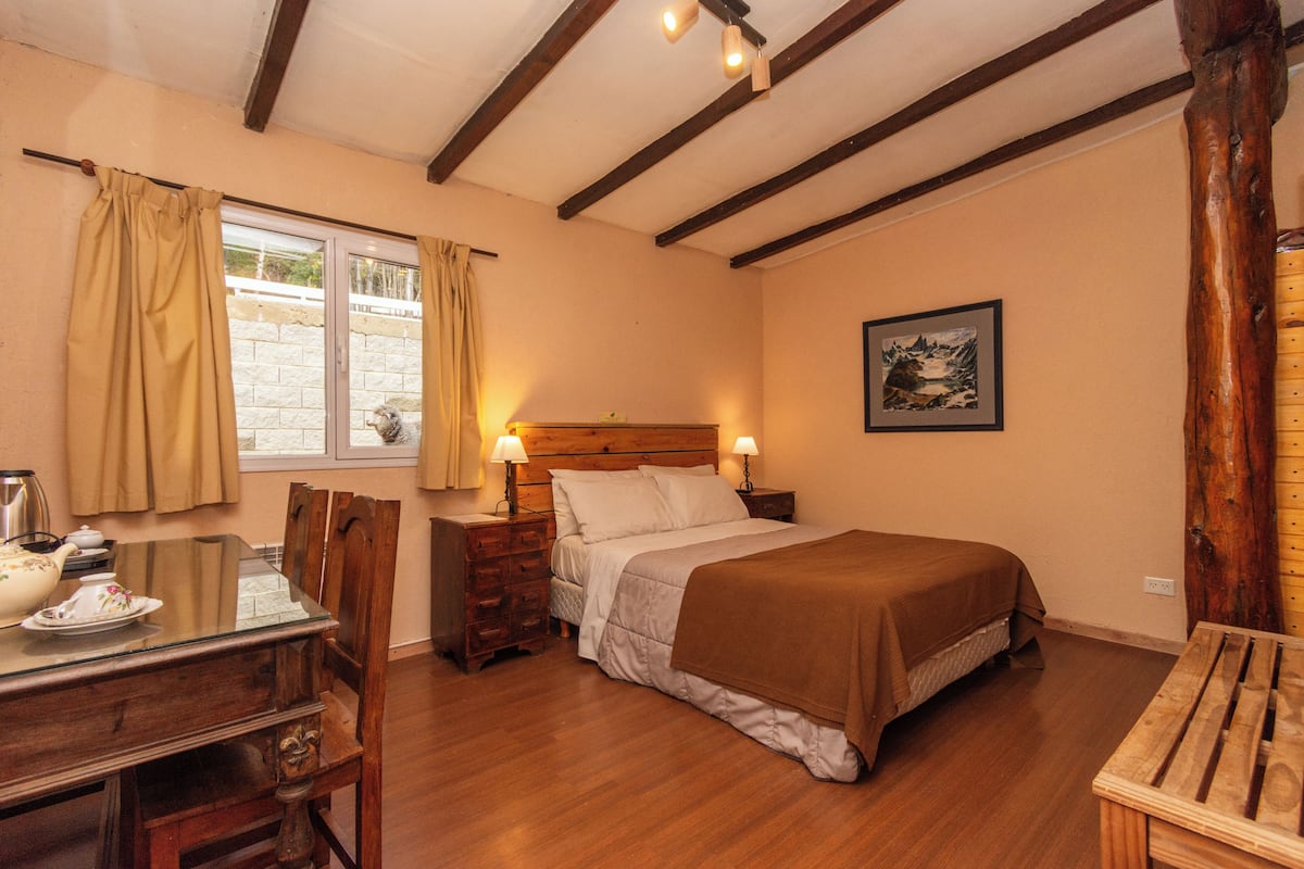 A bedroom with a wooden bed, bedside tables, a window with curtains, a dining table with chairs, and a painting on the wall.