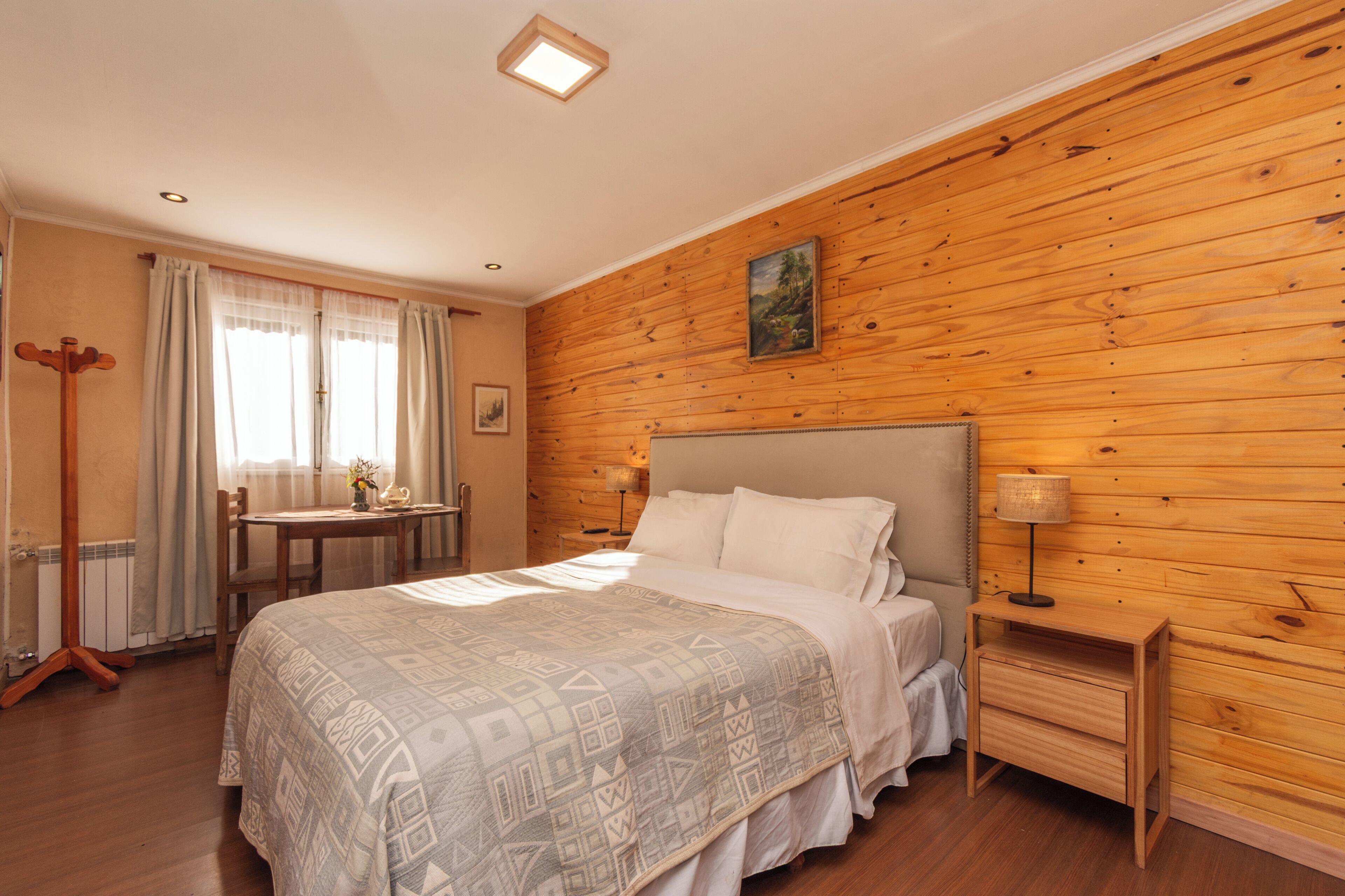 Un dormitorio con pared de madera, una cama, una mesita de noche, una lámpara, una silla, una mesa y una ventana con cortinas.